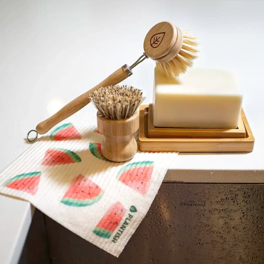 Cleaning supplies including a dish brush, scrubber, soap, and a cloth with watermelon design on a kitchen counter.