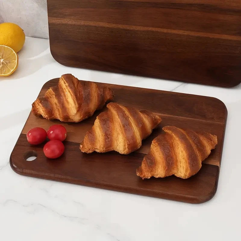 Three croissants on a wooden cutting board with two lemons and three tomatoes on a white surface.
