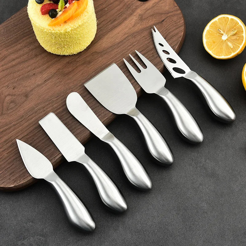 Set of silver cheese knives with black handles on a dark surface with a wooden board and fruit.