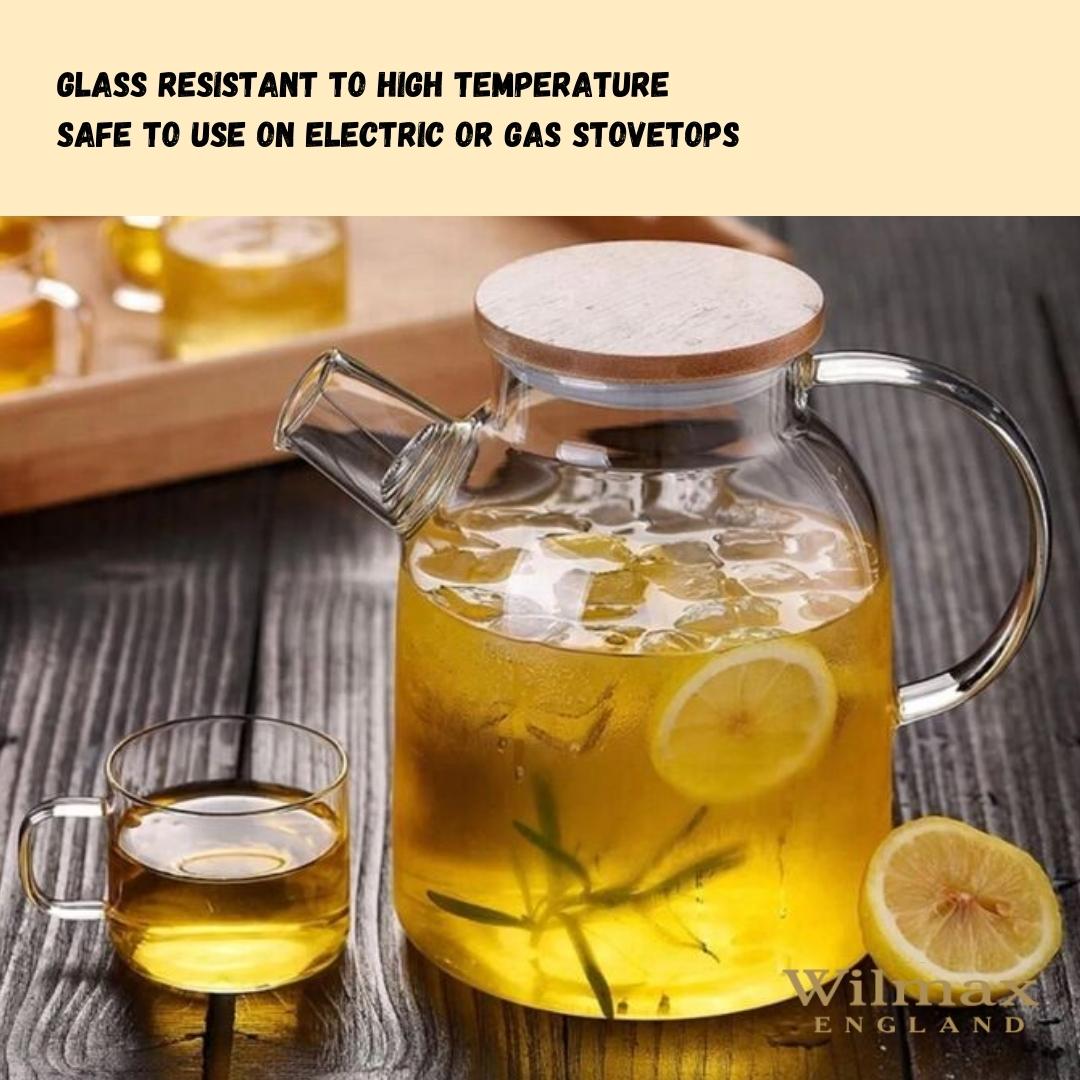 A clear glass teapot with a bamboo lid and stainless steel infuser, filled with golden-colored tea. The teapot has an ergonomic handle and is sitting on a wooden surface next to fresh tea leaves and a cup of tea.