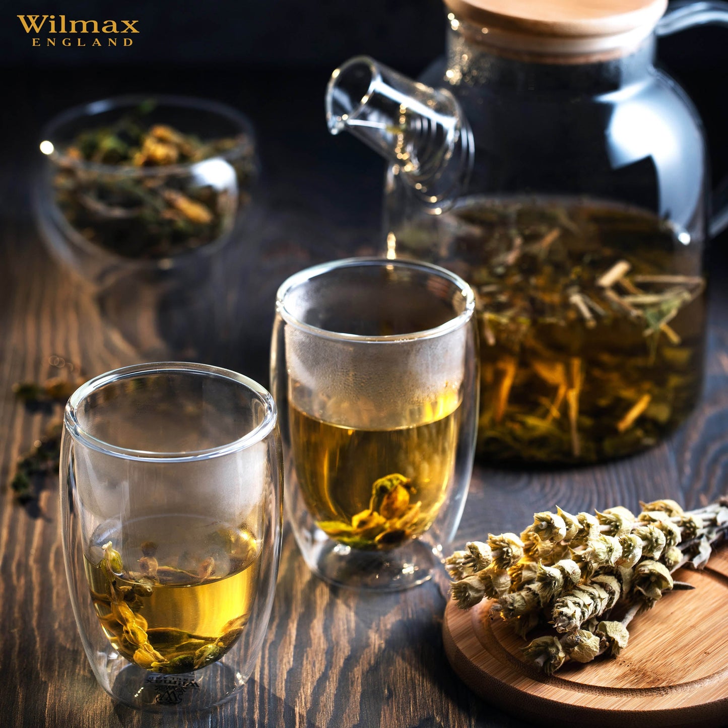 A clear glass teapot with a bamboo lid and stainless steel infuser, filled with golden-colored tea. The teapot has an ergonomic handle and is sitting on a wooden surface next to fresh tea leaves and a cup of tea.