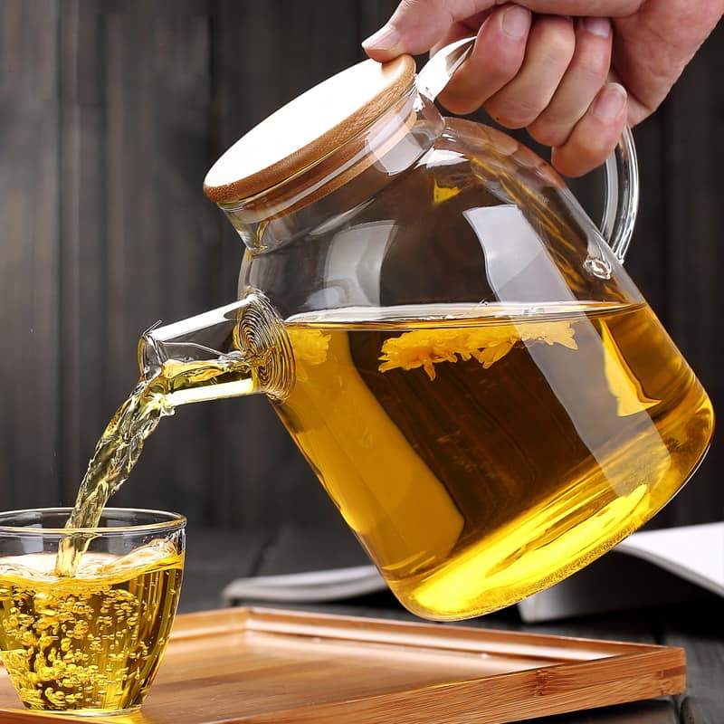 A clear glass teapot with a bamboo lid and stainless steel infuser, filled with golden-colored tea. The teapot has an ergonomic handle and is sitting on a wooden surface next to fresh tea leaves and a cup of tea.