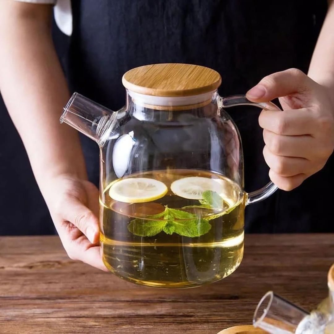 A clear glass teapot with a bamboo lid and stainless steel infuser, filled with golden-colored tea. The teapot has an ergonomic handle and is sitting on a wooden surface next to fresh tea leaves and a cup of tea.