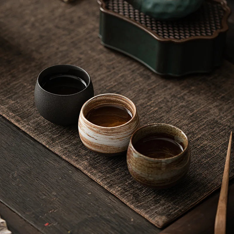 Three ceramic tea cups on a wooden surface with a teapot in the background.