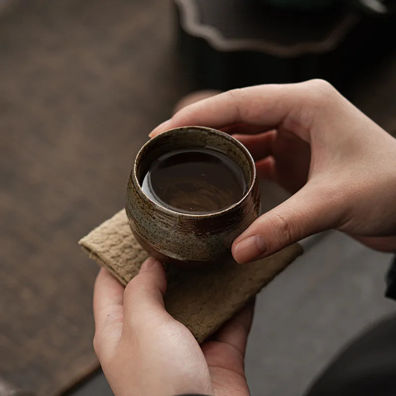 Person holding a small brown ceramic cup with a dark liquid on a wooden surface.