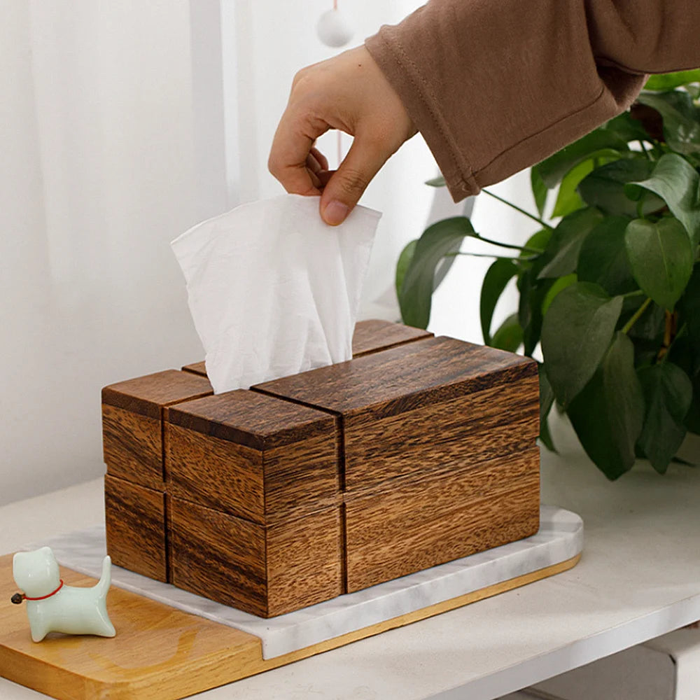 Wooden tissue box with a hand reaching in to pull out a tissue, placed on a marble surface with a plant in the background.