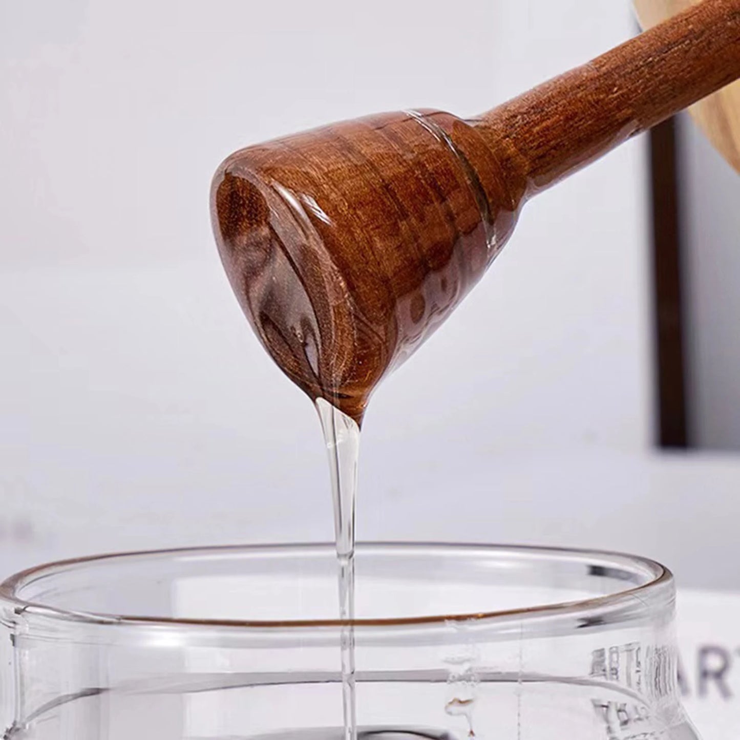 Wooden honey dipper pouring honey into a glass container with a blurred background
