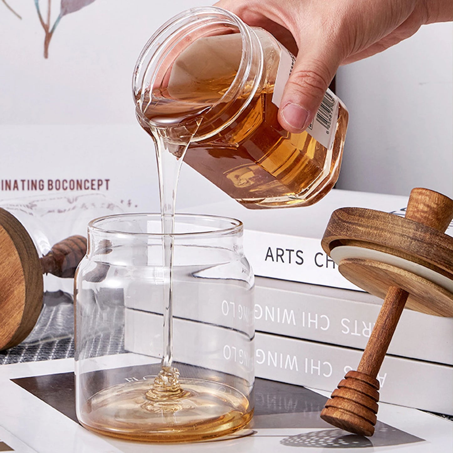 Hand pouring honey from a jar into a glass with a wooden honey dipper on a white surface.
