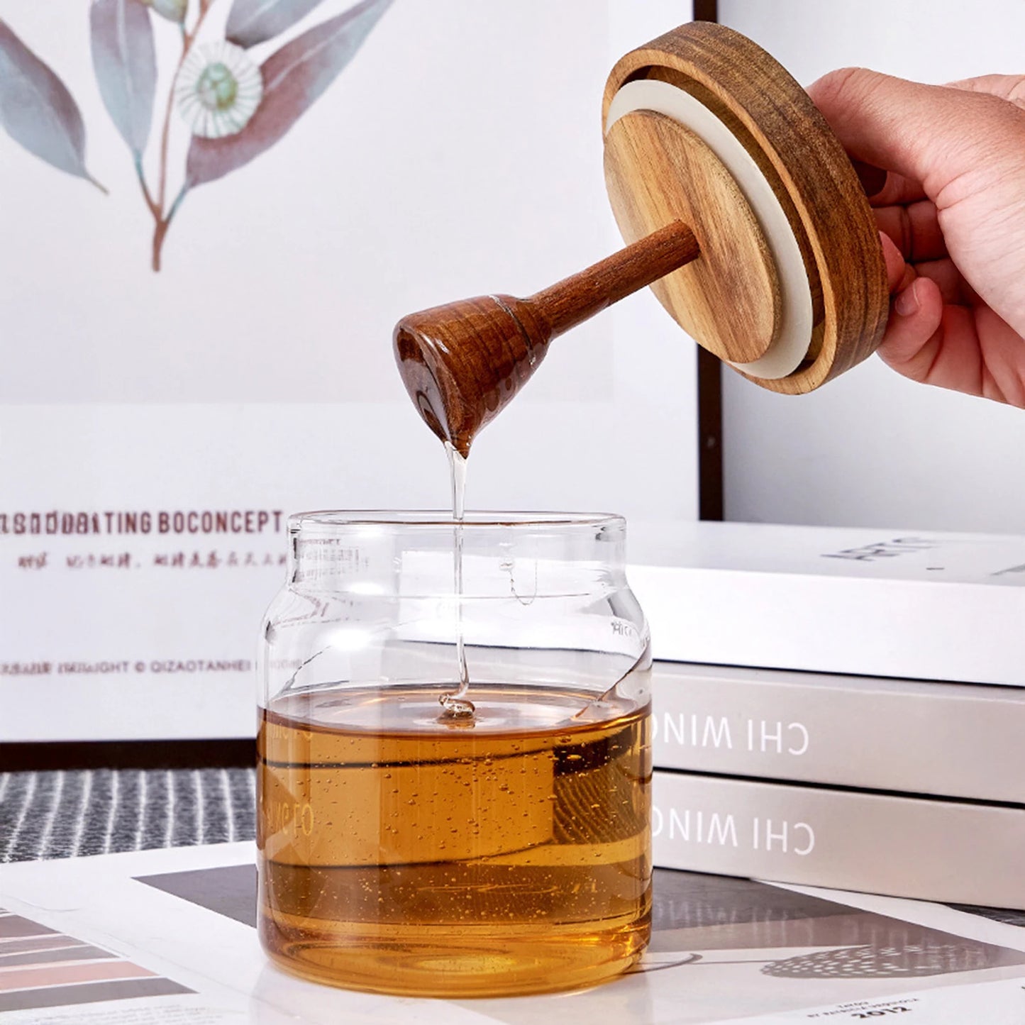 Hand pouring honey from a wooden honey dipper into a glass jar on a white surface with books in the background.