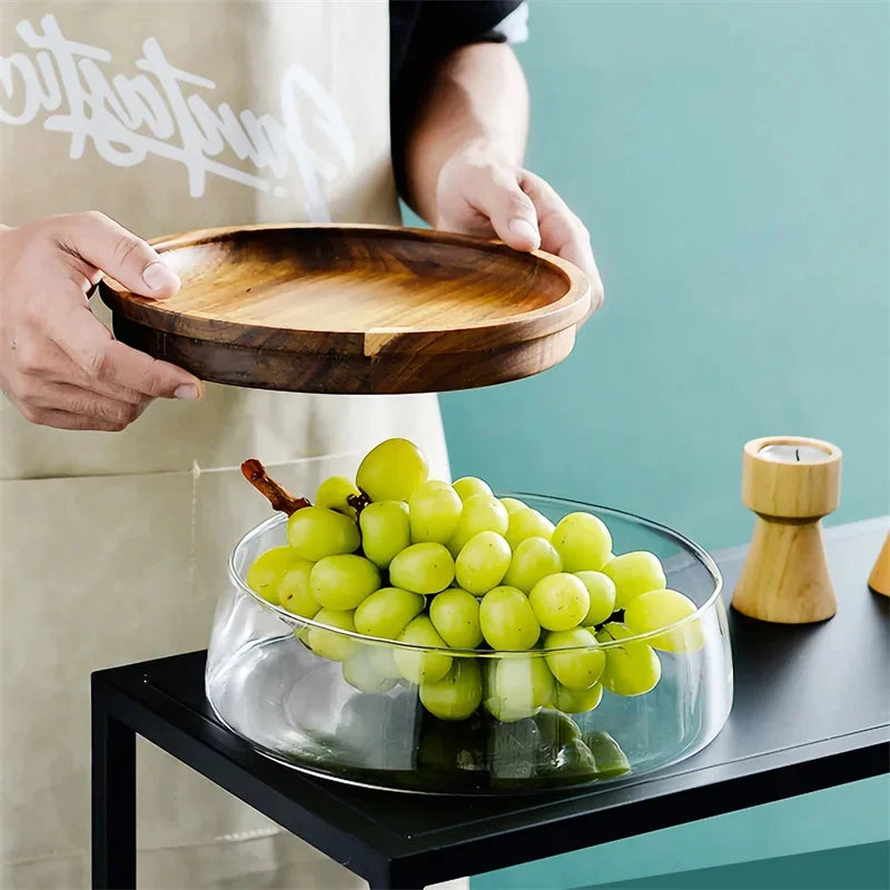 Person holding a wooden tray above a glass bowl with green grapes on a table.