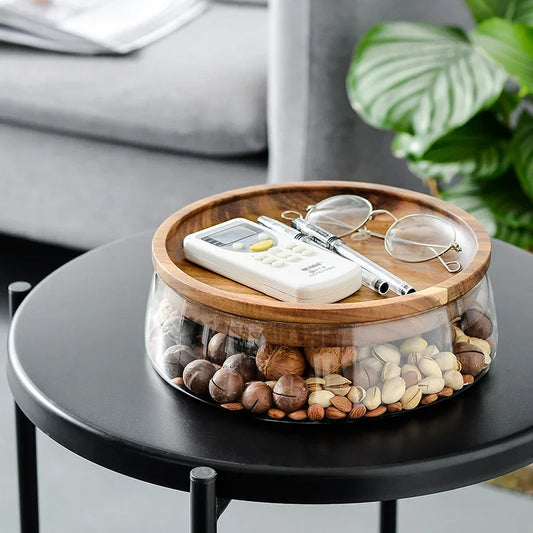 Decorative glass bowl with wooden lid on a black table, containing pebbles and small items.