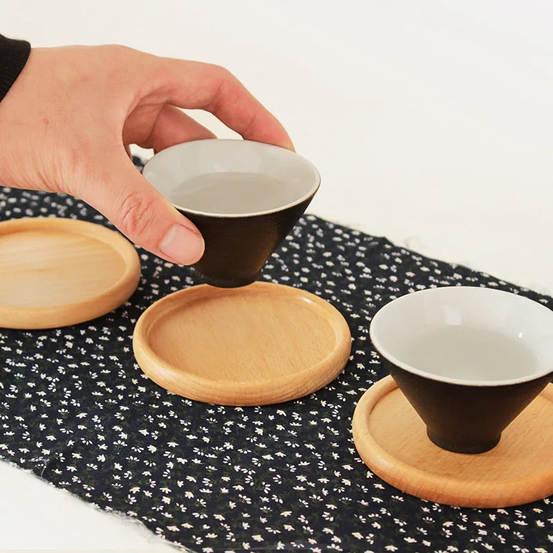 Person holding a small black cup with a white interior over wooden coasters on a black floral tablecloth.
