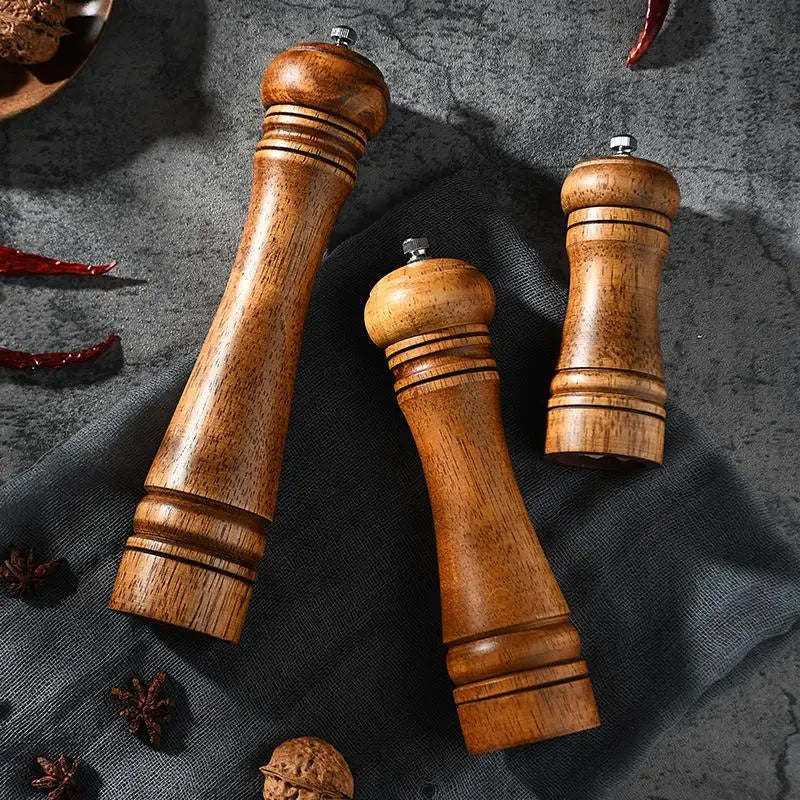 Three wooden pepper mills on a dark surface with spices and red peppers.