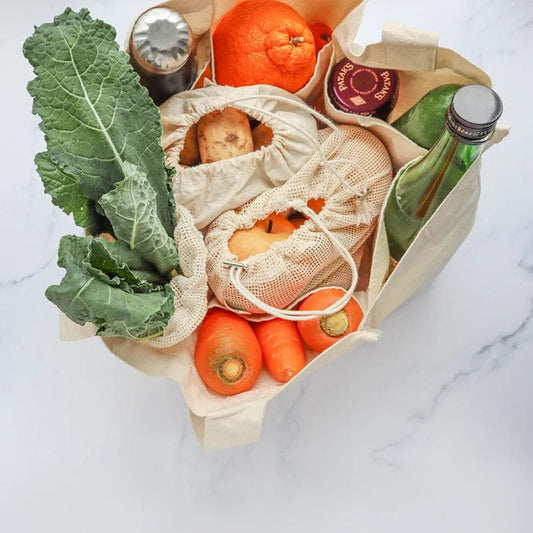 Open beige reusable grocery bag filled with fruits, vegetables, and bottles on a marble surface.