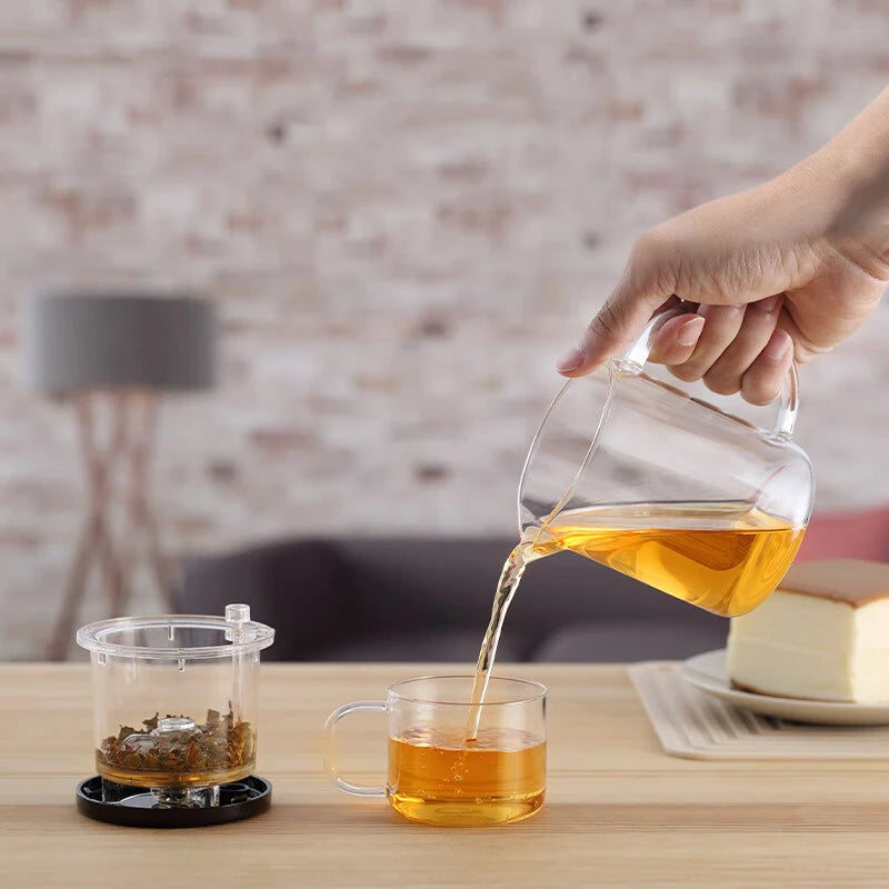 Person pouring tea from a glass teapot into a clear mug on a wooden table.