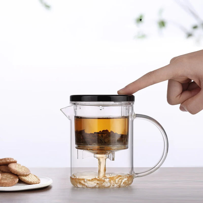 Clear glass teapot with black lid being held by a hand, filled with tea, on a light surface.