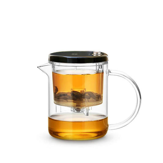 Clear glass teapot with black lid and handle, filled with tea on a white background