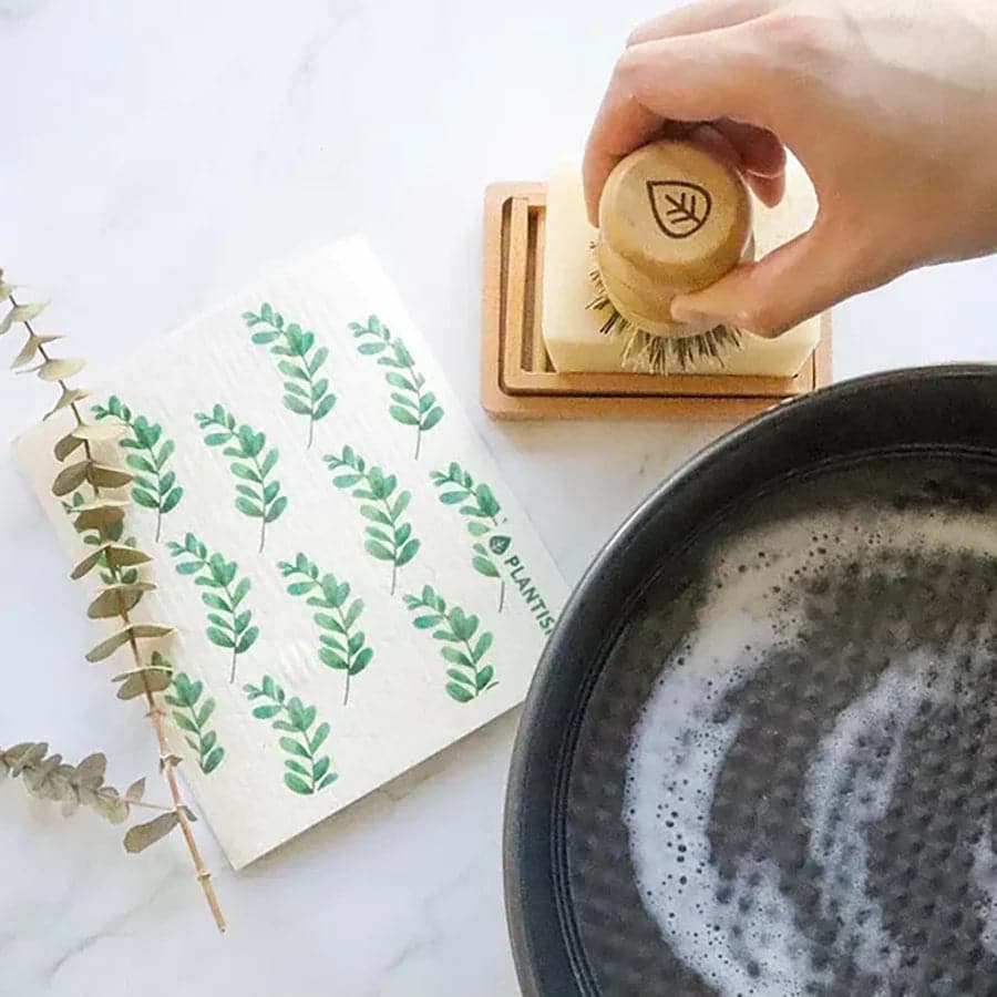 Hand using a wooden brush to clean a black pan with a white napkin featuring green leaf patterns on a white surface.